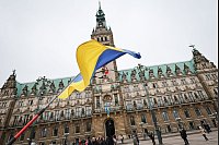 Ukraine-Krieg - Dritter Jahrestag. Ein Mann steht mit ukrainischer Fahne und Trauerflor bei der Gedenkminute „Hamburg steht still“ vor dem Rathaus. - © Christian Charisius/dpa