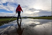 Eine Frau geht am vierten Advent mit Nordic Walking Stöcken durch eine Regenpfütze, in der sich der Himmel widerspiegelt. - © Thomas Warnack/dpa
