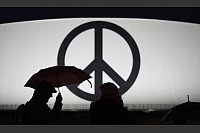 Passanten gehen mit Regenschirmen vorbei an einem leuchtenden Peace-Zeichen im strömenden Regen zum Musical-Theater «Stage Theater an der Elbe» im Hafen. - © Marcus Brandt/dpa