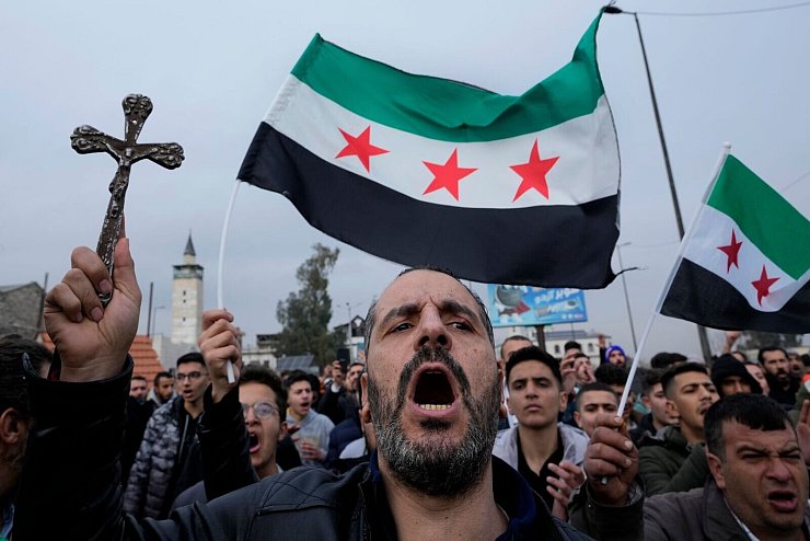 Ein syrischer Christ hält ein Kreuz hoch und ruft Parolen bei einem Protestmarsch in Damaskus, nachdem am Montagabend Unbekannte in Al-Sukailabija in der syrischen Provinz Hama den Weihnachtsbaum in Brand gesetzt hatten. - © Hussein Malla/AP/dpa