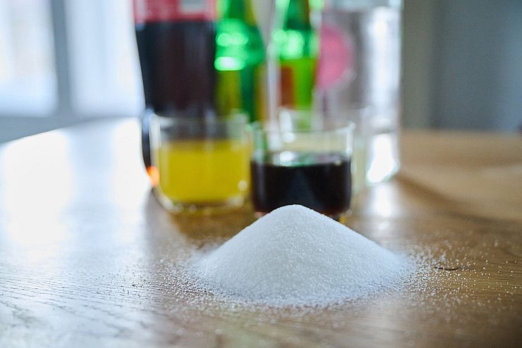 Drei unterschiedliche Arten Softdrinks und die Flaschen dazu stehen auf einem Tisch, während ein Berg von Zucker davor aufgeschüttet ist - © Annette Riedl/dpa