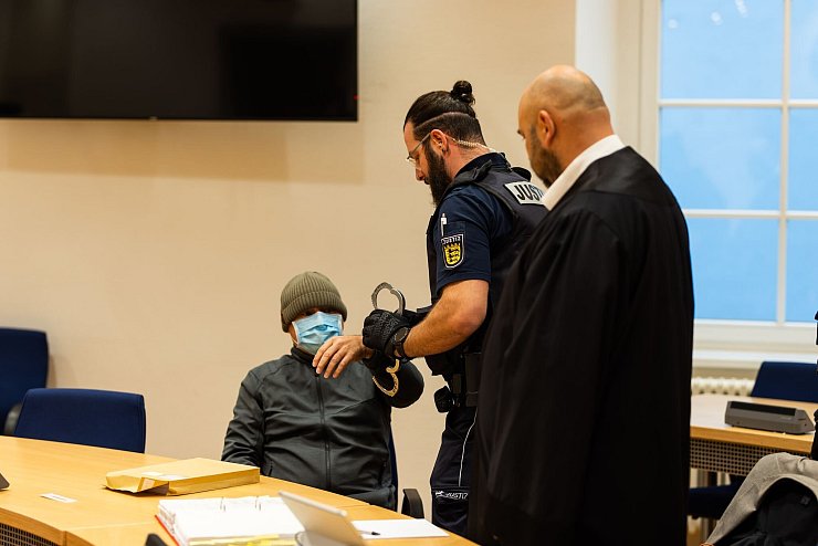 Ein Justizbeamter nimmt dem Angeklagten (links) im Gerichtssaal die Handschellen ab, während dessen Verteidiger daneben steht. - © Philipp von Ditfurth/dpa