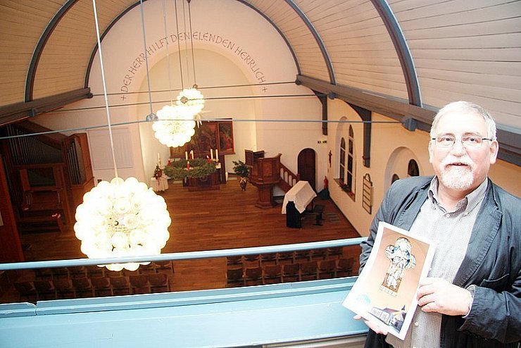 Pfarrer Ernst-August Korf hält ein Foto von dem Bild mit dem Jesusfenster in der Hand, das heute an der Westseite zu finden ist. Im Hintergrund ist der Hauptsaal der Kapelle mit dem Wandelaltar und der Kanzel zu erkennen - © 