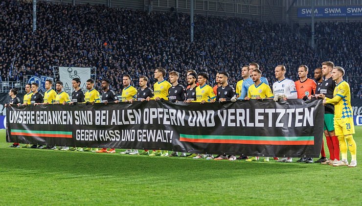 Beide Teams stehen während einer Schweigeminute zusammen hinter einem Transparent mit der Aufschrift: «Unsere Gedanken sind mit allen Opfern und Verletzten - gegen Hass und Gewalt!». - © Andreas Gora/dpa