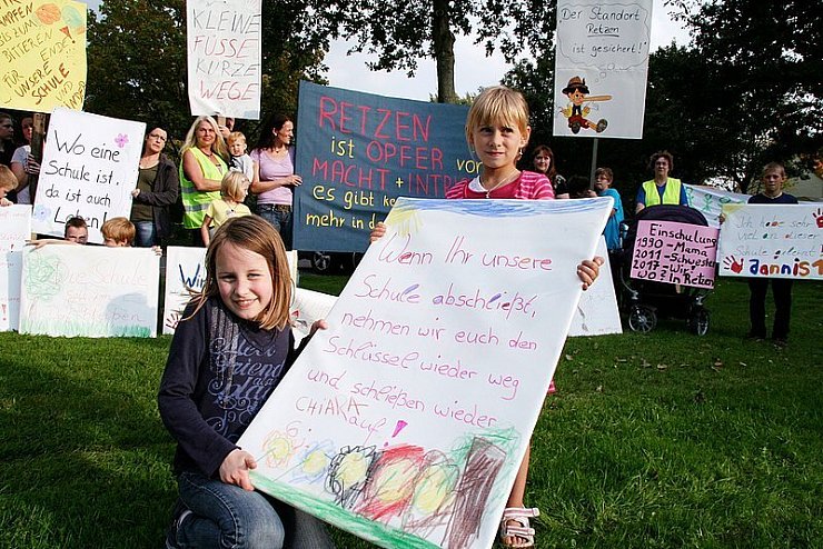 Ein kindlicher Wunsch: "Wenn Ihr unsere Schule abschließt, nehmen wir euch den Schlüssel wieder weg und schließen wieder auf", hat Chiara (rechts) auf ihr Plakat geschrieben, das sie zusammen mit Anne hält. Im Hintergrund stehen die Retzer Eltern und Kinder mit weiteren Plakaten und Transparenten vor ihrem Protestmarsch durch das Dorf. - ©