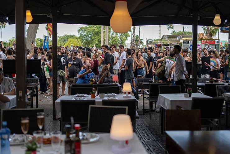 Ein Mann und eine Frau auf der Terrasse eines Restaurants in Barcelona lassen sich von vorbeiziehenden Demonstranten gegen den Massentourismus offenbar nicht aus der Ruhe bringen. - © Lorena Sopêna/EUROPA PRESS/dpa