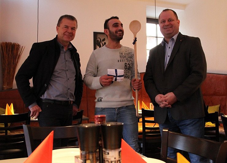 Arne Brand, Allg. Vertreter der Verbandsvorsteherin/des Verbandsvorstehers, und Jürgen Hoppe überreichen Suar Kassem einen Kochlöffel sowie eine Kochmütze und gratulieren zur Eröffnung von „El Molino".