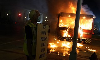 Ein Demonstrant steht mit einem Transparentvor einem brennenden Müllwagen. - Foto: David Goldman/AP/dpa