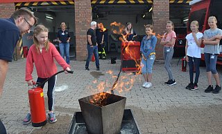 Emma löscht unter Anleitung von Patrick Althof das Feuer in der Schale, die anderen Kinder der Ferienspiel-Aktion bei der Feuerwehr schauen zu. - Sibylle Kemna Emma löscht unter Anleitung von Patrick Althof das Feuer in der Schale, die anderen Kinder der Ferienspiel-Aktion bei der Feuerwehr schauen zu. - Sibylle Kemna