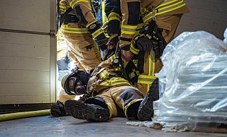 Der bewusstlose Feuerwehrmann Marcel Knorr wird vom Sicherheitstrupp und seinem Trupppartner aus der Lagerhalle evakuiert und an die weiteren Kräfte vor der Halle übergeben. - FREITAG-TV