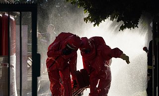 Feuerwehrleute in chemischen Schutzanzügen retten einen Verletzten, während im Hintergrund die giftigen Gase mit Wasser niedergeschlagen werden. - Freitag-TV