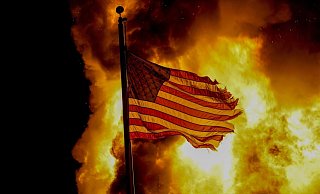 Eine US-Flagge weht vor einem Feuer, das bei Protesten vor dem Gebäude einer Strafvollzugsanstalt in Kenosha lodert. - Foto: Morry Gash/AP/dpa