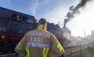 Stolz winkt der Lokführer den amüsierten Feuerwehrleuten aus Lage zu. - Freitag TV/Stock