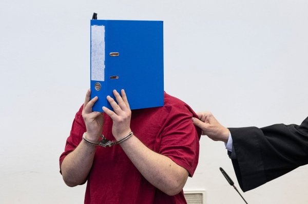 Der Angeklagte steht hinter der Anklagebank und verdeckt sein Gesicht hinter einem Aktenordner. - © Boris Roessler/dpa