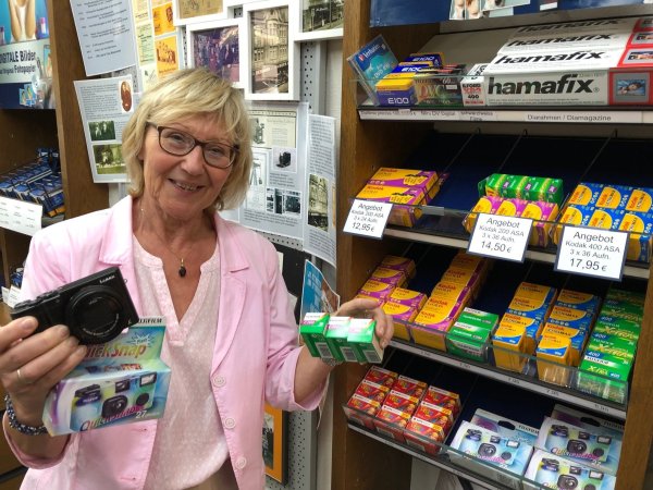 Fotogeschäft im Wandel: Laut Ingrid Mildner-Biesemeier wird es immer schwieriger, analoge Filme zu bekommen, wie sie sie in der Hand hält und von denen sich weitere im Regal hinter ihr befinden. "Schuld" daran ist die rasante Entwicklung der digitalen Fotografie. Ein aktuelles Panasonic-Modell hält sie in der anderen Hand - neben einer analogen Einwegkamera, die ebenfalls aktuell in Mode ist. - © Thomas Reineke