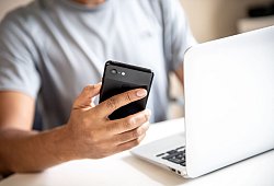 Mann hält vor einem Laptop sein Smartphone in der Hand - Zacharie Scheurer/dpa-tmn