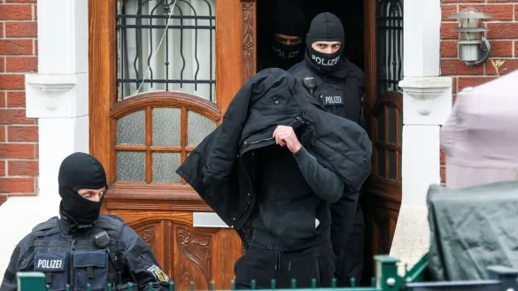 Polizeibeamte führen einen Tatverdächtigen aus einem durchsuchten Haus. - © Christoph Reichwein/dpa