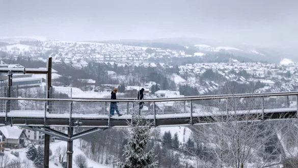 Spaziergänger gehen eine Brücke neben der Winterberger Bobbahn mit Blick auf die Landschaft im Schnee. - © David Inderlied/dpa