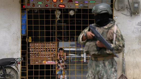 Ein Junge schaut hinter den Gitterstäben eines geöffneten Elektronikgeschäfts hervor, während ein Soldat an einem von der Armee eingerichteten Sicherheitskontrollpunkt Wache steht. - © Martin Mejia/AP/dpa
