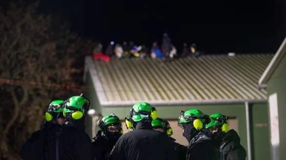 Klimaaktivisten sitzen auf einem Scheunendach, im Vordergrund stehen Höhenkletterer der Polizei zu einer Lagebesprechung zusammen. - © Thomas Banneyer/dpa/Archivbild