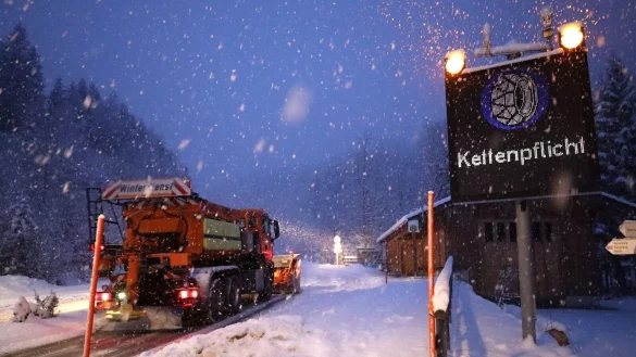 Ein Schild weist auf die Schneekettenpflicht am Riedbergpass im bayerischen Landkreis Oberallgäu hin, während daneben ein Räumfahrzeug den Schnee von der Straße räumt. - © Davor Knappmeyer/dpa