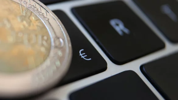 Eine Zwei-Euro-M&uuml;nze liegt auf der Tastatur eines Laptops neben einem Eurozeichen. - &copy; Karl-Josef Hildenbrand/dpa