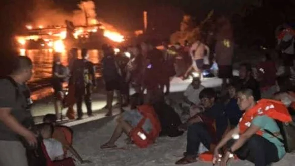 Nach ihrer Rettung ruhen sich Überlebende an Land aus. Im Hintergrund ist die brennende Fähre zu sehen. - © Uncredited/PHILIPPINE COAST GUARD/AP/dpa