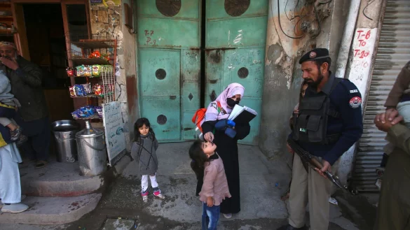 Ein Polizist in Pakistan steht Wache, w&auml;hrend einem Kind ein Polio-Impfstoff verabreicht wird. - &copy; Muhammad Sajjad/AP/dpa