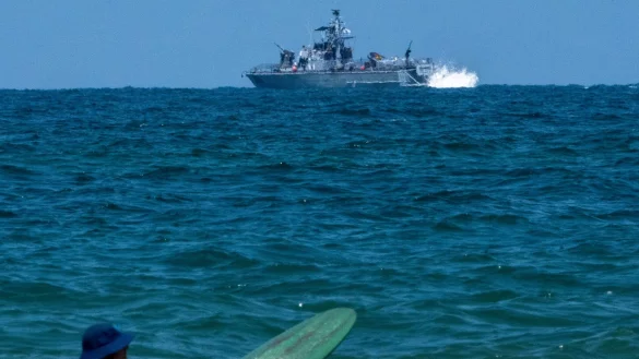 Ein Surfer wartet auf eine Welle, w&auml;hrend ein israelisches Marineschiff im Mittelmeer vor der K&uuml;ste von Hadera patrouilliert. - &copy; Ariel Schalit/AP