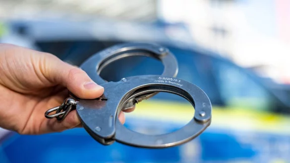 Eine Hand hält Handschellen vor einen Streifenwagen der Polizei. - © David Inderlied/dpa/Illustration