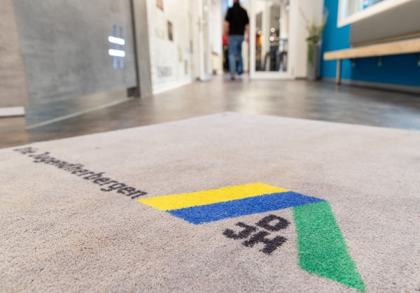 Blick auf den Teppich am Eingang einer Jugendherberge mit Logo des Deutschen Jugendherbergswerks (DJH). - © Friso Gentsch/dpa/Archivbild