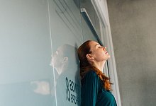 Frau lehnt erschöpft an einer Glaswand im Büro - Zacharie Scheurer/dpa-tmn