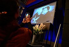Eine Frau fotografiert mit ihrem Smartphone die B&uuml;hne mit Blumenstrau&szlig; und einem Foto des Verstorbenen auf einer Leinwand. - Jonas Walzberg/dpa