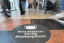 Passanten laufen durch ein Einkaufszentrum auf dessen Fu&szlig;boden ein Hinweisschild zur Maskenpflicht aufgeklebt ist. - Danny Gohlke/dpa