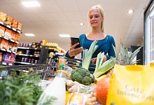 Frau mit Einkaufswagen schaut aufs Handy - Benjamin Nolte/dpa-tmn