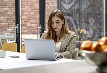 A woman is focused on her work at a laptop on a white table, with a smartphone and oranges nearby, in a well-lit room with windows. - picture alliance / Image Source