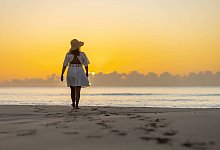 Back view of an unrecognizable woman enjoying a peaceful walk along the shore at sunrise, with the warm glow of the sun - IMAGO/Addictive Stock
