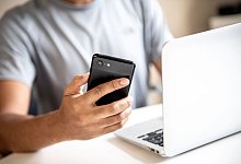 Mann h&auml;lt ein Handy in der Hand und sitzt am Laptop. - Zacharie Scheurer/dpa-tmn