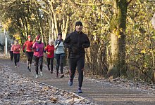 A group of people jogging on a path covered with fallen leaves in a forest, wearing colorful athletic clothing. Sale Water Park, Manchester, UK - picture alliance