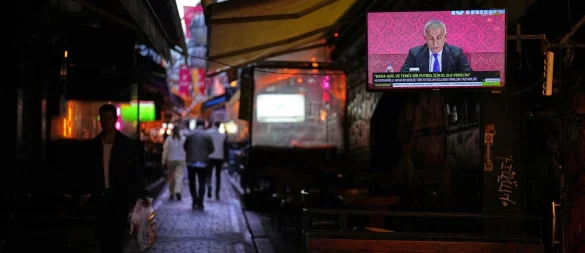 Menschen gehen an einem Fernsehbildschirm vorbei, auf dem der Präsident des Türkischen Fußballverbands, Ibrahim Haciosmanoglu, während einer Pressekonferenz zu sehen ist. - © Francisco Seco/AP/dpa