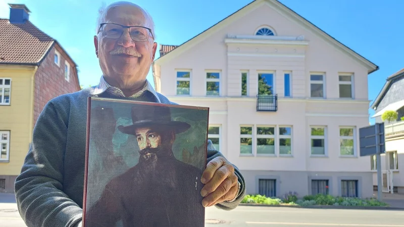 Dr. Hans-Joachim Keil präsentiert die Abbildung eines Porträts von Adolf Neumann-Hofer vor dessen ehemaligen Wohnhaus in der Leopoldstraße. Das Gemälde stammt vom Künstler Bruno Wittenstein (1876-1968), ist um 1920 mit Öl auf Leinwand entstanden und zeigt Adolf Neumann-Hofer mit seinem Markenzeichen: dem langen, dunklen Bart. - © Carolin Brokmann-Förster