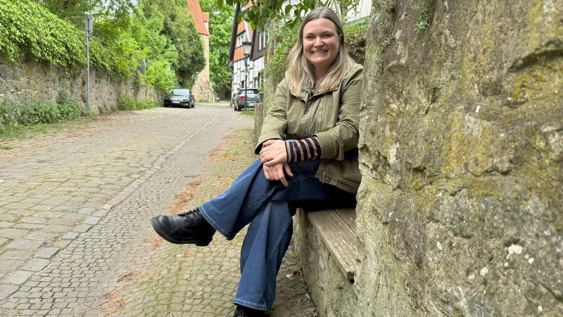 Sonja Beinlich sitzt auf einer Bank, die in einer Mauer in der Turmstraße eingelassen ist. Auf der gegenüberliegenden Seite befinden sich die Stadtmauer und im Hintergrund der Katzenturm. - © Sven Kienscherf