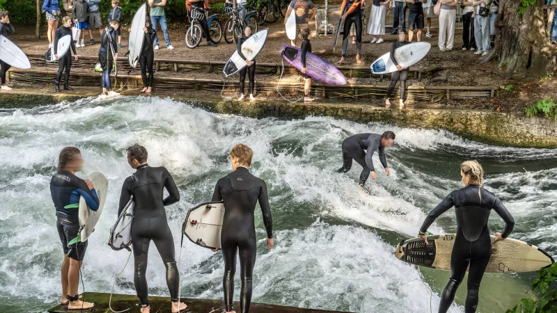 Surfer stehen am Eisbach in M&uuml;nchen. Zuschauer beobachten gespannt das Geschehen. - &copy; IMAGO/Wolfgang Maria Weber
