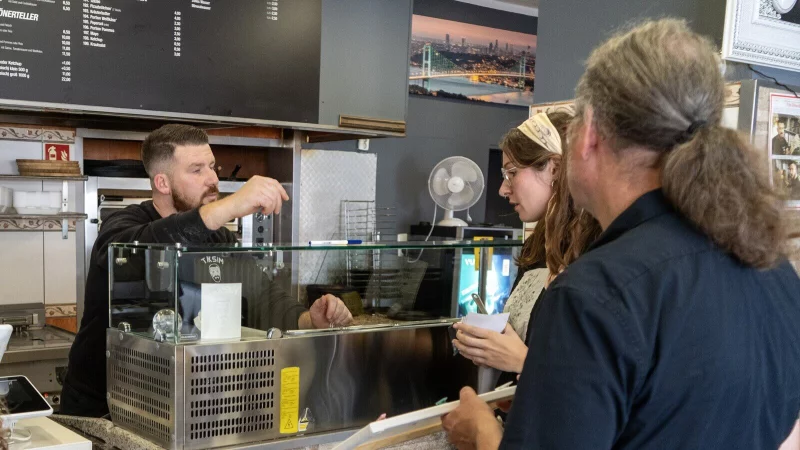 Matthias Zizelmann (rechts) und Mona Lorenz von HBM Chribal &uuml;bergeben Tafel und Plakate f&uuml;r die Aktion Miteinander, F&uuml;reinander an Mesut Ocak von Taksim Pizza & D&ouml;ner. - &copy; Stadt Horn-Bad Meinberg