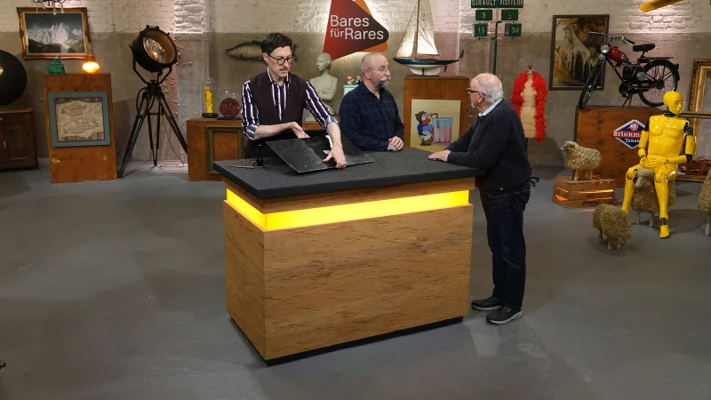 Experte Colmar Schulte-Goltz (v.l.) begutachtet bei "Bares f&uuml;r Rares" das Miele-Emailleschild, w&auml;hrend Moderator Horst Lichter mit dem Harsewinkeler Georg Terlutter redet. - &copy; ZDF/Warner