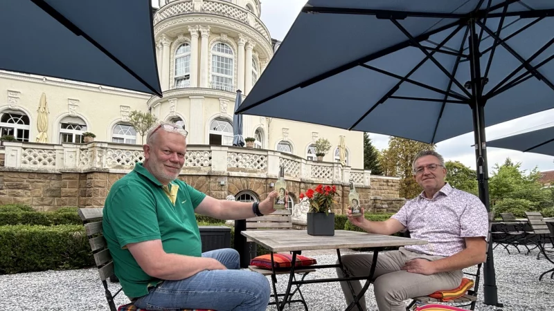 Jupp Westfeld, Cheforganisator der „Sommer-Radl-Sause“ (links), und Andreas Gerbig, Direktor der "Villa Dürkopp", stoßen mit einer Salzufler Sole-Limo auf die „Sommer-Radl-Sause“ an, die am 7. September auch in dem Biergarten am Fuß der Gründerzeit-Fassade Station macht. - © Thomas Reineke
