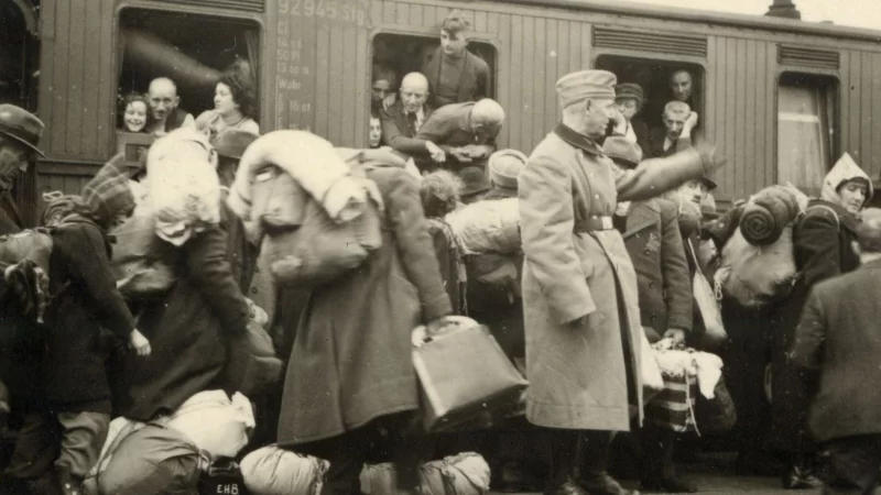 Jüdinnen und Juden am Bielefelder Hauptbahnhof auf dem Weg zu den ihnen zugewiesenen Waggons. Im Zug sitzen bereits Deportierte aus dem Kreis Münster. Im Vordergrund steht ein Mitglied des Begleitkommandos mit erhobener Hand. - © Stadtarchiv Bielefeld