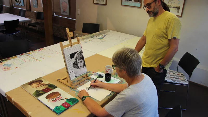 Sigrid Rutz malt ein Porträt mit Feder und Tinte. Igor Oster, Leiter der Kunstwerkstatt Eben-Ezer, schaut ihr über die Schulter. Im Hintergrund sind Bilder der Teilnehmer zu sehen. - © Nadine Uphoff