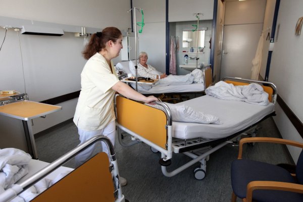 Eine Kliniksericekraft schiebt ein Bett aus einem Patientenzimmer auf der Station 3a. Im Hintergrund ist die Waschnische zu sehen, die sich die Zimmergemeinschaft teilen muss. Die Toilette ist auf dem Flur. - © Bernhard Preuß