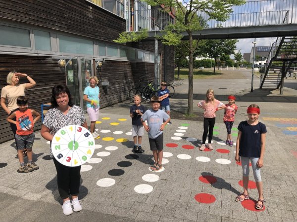 Anke Brand (im Vordergrund mit einer "Würfel"-Scheibe) hat mit OGS-Kindern aus Knetterheide in den Sommerferien ein lebendiges "Mensch-Ärgere-Dich-nicht-Spiel" auf dem Pausenhof aufgebracht. - © Thomas Reineke